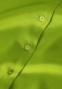 Helle grüne Satin-Stoff mit glatter Textur, versehen mit zwei hellgrünen Knöpfen, von denen einer sichtbar an einer Falte angenäht ist.