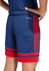 Marineblaue Sportshorts mit roten seitlichen Streifen, ausgestattet mit einem Kordelzugbund und strukturiertem Stoff. "AEROREADY"-Logo auf der Rückseite gedruckt.