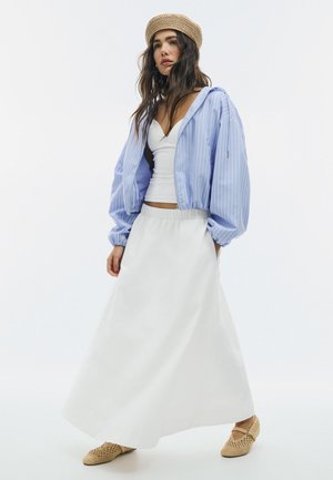 Ženska v modrem črtastem suknjiču, beli majici, dolgi beli krilu, bež baretki in tkanih bež balerinkah, stoji pred enobarvnim ozadjem.