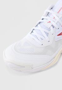 Κοντινό πλάνο λευκού αθλητικού παπουτσιού με ύφασμα από δίχτυ, λευκά κορδόνια και κόκκινη λεπτομέρεια λογότυπου στο πλάι και τη γλώσσα.