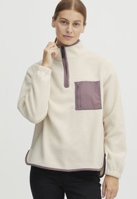 Pulover od flisa u krem boji, s visokim ovratnikom, polu-zip zatvaračem i bordo džepom. Teksturirana tkanina s kontrastnim detaljima.