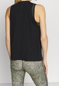 Kvinna som bär en svart ärmlös topp över en randig sportbehå med leggings i leopardmönster, sedd bakifrån mot en enkel bakgrund.