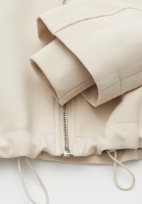 Beige athletic jacket featuring a zip closure, ribbed cuffs, and adjustable hem with drawstrings. Smooth fabric with a matte finish.