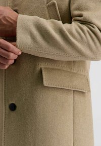 Beige Wollmantel mit glatter Textur, ausgestattet mit einem einzigen Knopf, einer Leistentasche und leicht umgeschlagenen Ärmelenden.