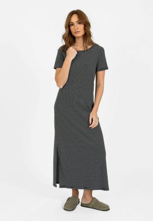 Femme debout portant une robe maxi rayée noir et blanc à manches courtes avec fente latérale et chaussures vertes à enfiler sur fond blanc.