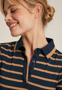 Navy- und Beige-gestreiftes Poloshirt mit einem geknöpften Kragen, gerippter Textur und auffälligen Nähten, getragen von einer Person im Seitenprofil.