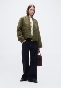 Γυναίκα που φοράει μία κυπαρισσί μπουφάν με καπιτονέ λεπτομέρειες πάνω από ένα λευκό μπλουζάκι με γραφικό σχέδιο, φαρδιά σκουρόχρωμα τζιν, μαύρα παπούτσια και κρατά μία καρό καφέ τσάντα χειρός.