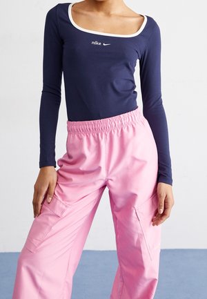 Person iført navyblå Nike langærmet top med hvide kanter og lyserøde cargobukser, stående mod en ensfarvet baggrund.