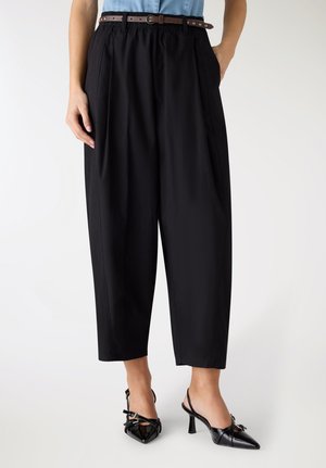 Donna che indossa pantaloni neri a gamba larga tagliati sopra la caviglia con una cintura marrone, camicia azzurro chiaro e décolleté slingback nere con punta affilata.