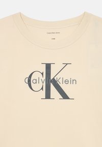 Béžové bavlněné tričko s kulatým výstřihem, na kterém je velké černé logo "CK" a pod ním stříbrně vytištěné "Calvin Klein".