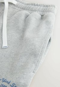 Lichtgrijze joggingbroek gemaakt van zachte, gestructureerde stof, met een witte trekkoord en afgedrukte blauwe tekst op de zijkant.