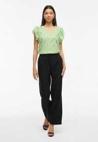 Blusa verde chiaro a V con maniche a volant e un motivo intricato, abbinata a pantaloni neri a gamba larga e sandali neri con tacco.