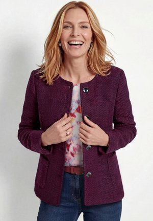 Vrouw met schouderlang blond haar die lacht, draagt een paarse getextureerde jas over een kleurrijk blouse en blauwe spijkerbroek.