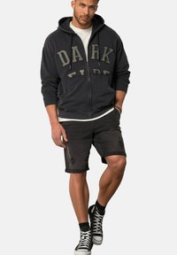Felpa nera con zip e testo "DARK SIDE", abbinata a pantaloni corti neri arrotolati e sneakers alte nere. Maglietta bianca sotto.