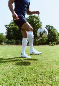 Des crampons blancs avec un logo doré, un short noir et des protège-tibias blancs ; dribblant un ballon de football noir et blanc sur de l'herbe verte.