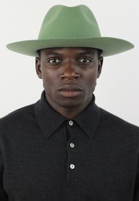 Grüner Filzhut mit breiter Krempe, getragen mit einem dunklen Poloshirt mit Kragen und Knopfleiste. Der Stoff wirkt strukturiert und eng anliegend.