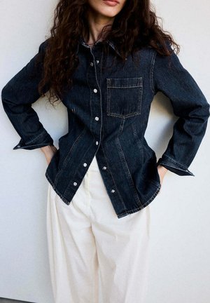 Femme portant une veste en jean foncée avec des boutons blancs et un pantalon crème à jambes larges, posant les mains dans les poches devant un fond uni.