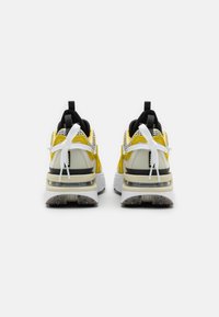 Pár žlutých a bílých tenisek s černými detaily, bílými šněrovacími tkaničkami, silnými podrážkami a poutky, viděný ze zadní strany na bílém pozadí.