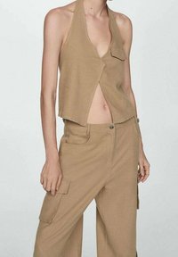 Beige haltertop met een V-hals en twee voorkruiszakken, gecombineerd met beige wijde broekspijpen met een zijzak en knoopsluiting.