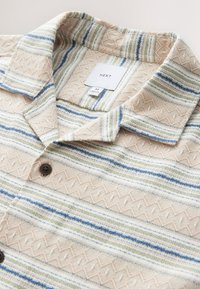 Camicia con colletto in tessuto beige con righe texturizzate in blu e verde. Bottoni marroni e etichetta appesa con informazioni sulla taglia.