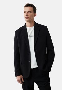 Sort blazer lavet af tekstureret stof, med fadeslag, to-knaps lukning og forlommer. Båret over en hvid T-shirt.