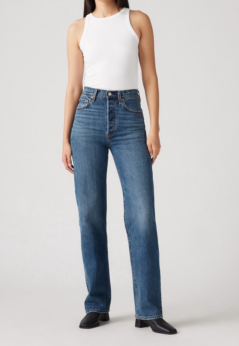 Femme portant un jean bleu taille haute à jambes droites, un top blanc sans manches en côtes, et des bottines noires à bout carré et talon, debout sur un fond uni.