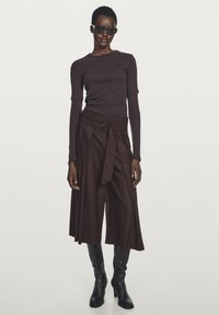 Bruine top met lange mouwen gecombineerd met wijde bruine broek met riem, aangevuld met zwarte kniehoge laarzen en zonnebril.