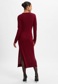 Burgunderfarbenes Kleid mit langen Ärmeln, taillierter Silhouette, seitlichem Schlitz und geripptem Material, kombiniert mit schwarzen High Heels.