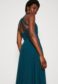 Rochie de seară teal cu un corset din dantelă ajustat și fustă fluidă. Are bretele încrucișate și detalii de dantelă intricate pe spate.