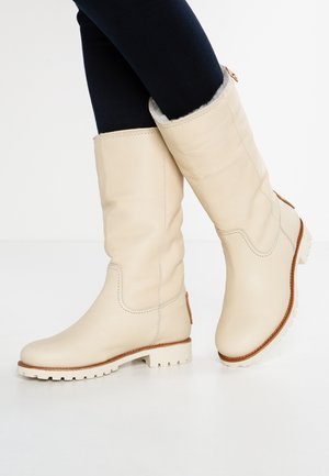 Bottes en cuir beige montant jusqu'aux genoux avec doublure en fourrure et semelles robustes, portées avec des leggings noirs, montrées croisées sur un fond blanc.