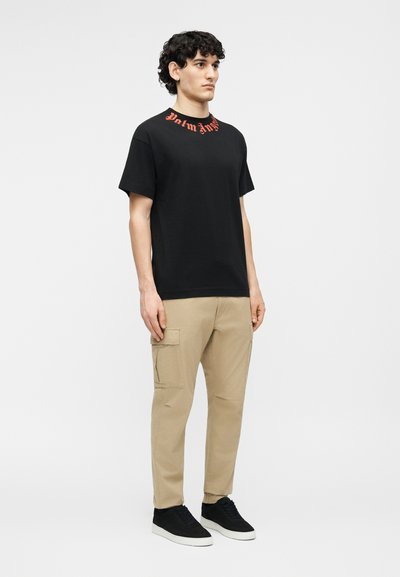 Fiatal férfi göndör hajjal, fekete pólót visel piros felirattal a gallérján, bézs cargo nadrágot és fekete sportcipőt, mozdulatlanul áll.