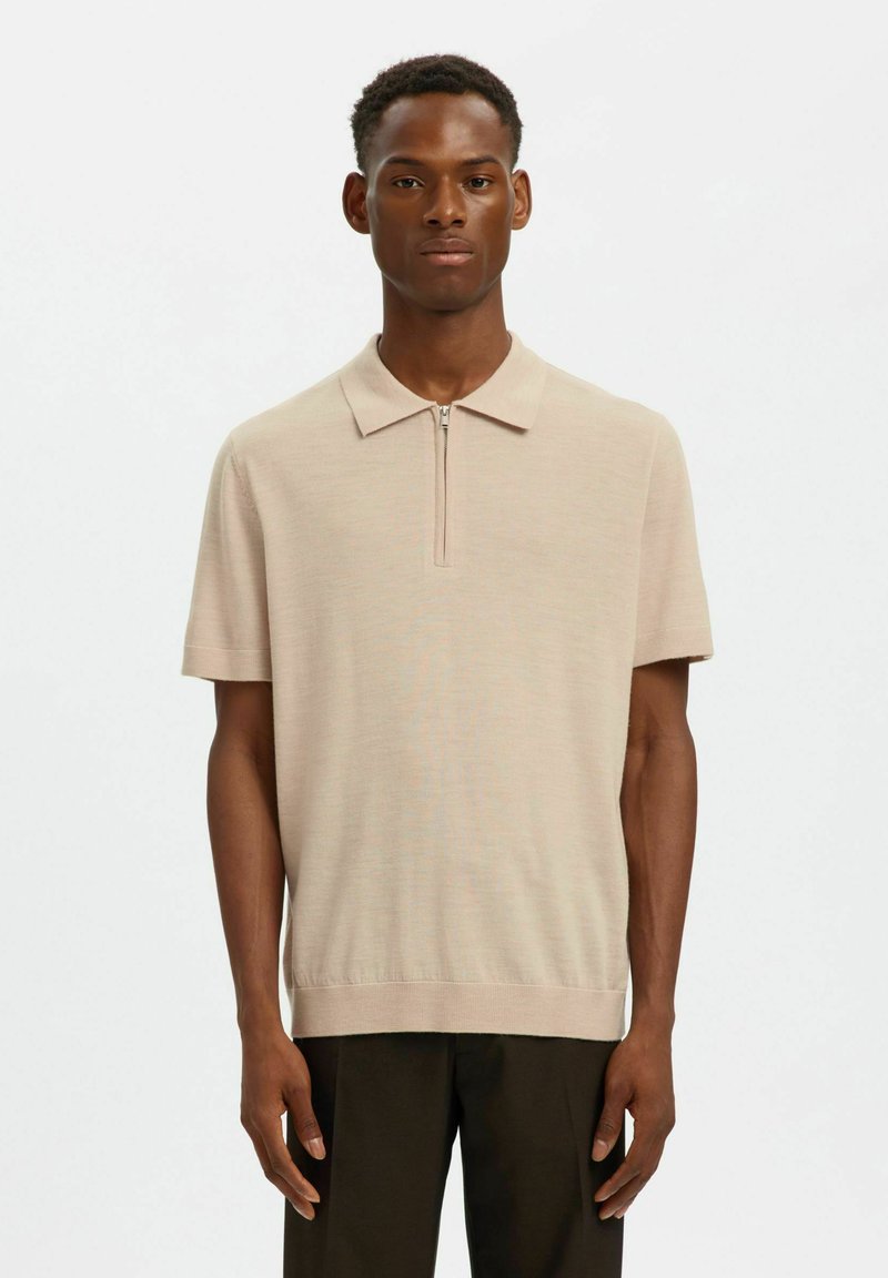 Homme portant un polo en maille beige à manches courtes avec un col à fermeture éclair et un pantalon foncé, debout devant un fond blanc uni.