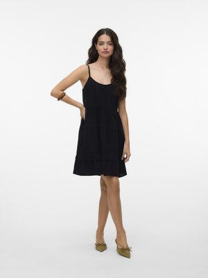 Femeie în picioare, cu o mână pe șold, purtând o rochie neagră fără mâneci, până la genunchi, și pantofi oliv cu vârf ascuțit și funda.