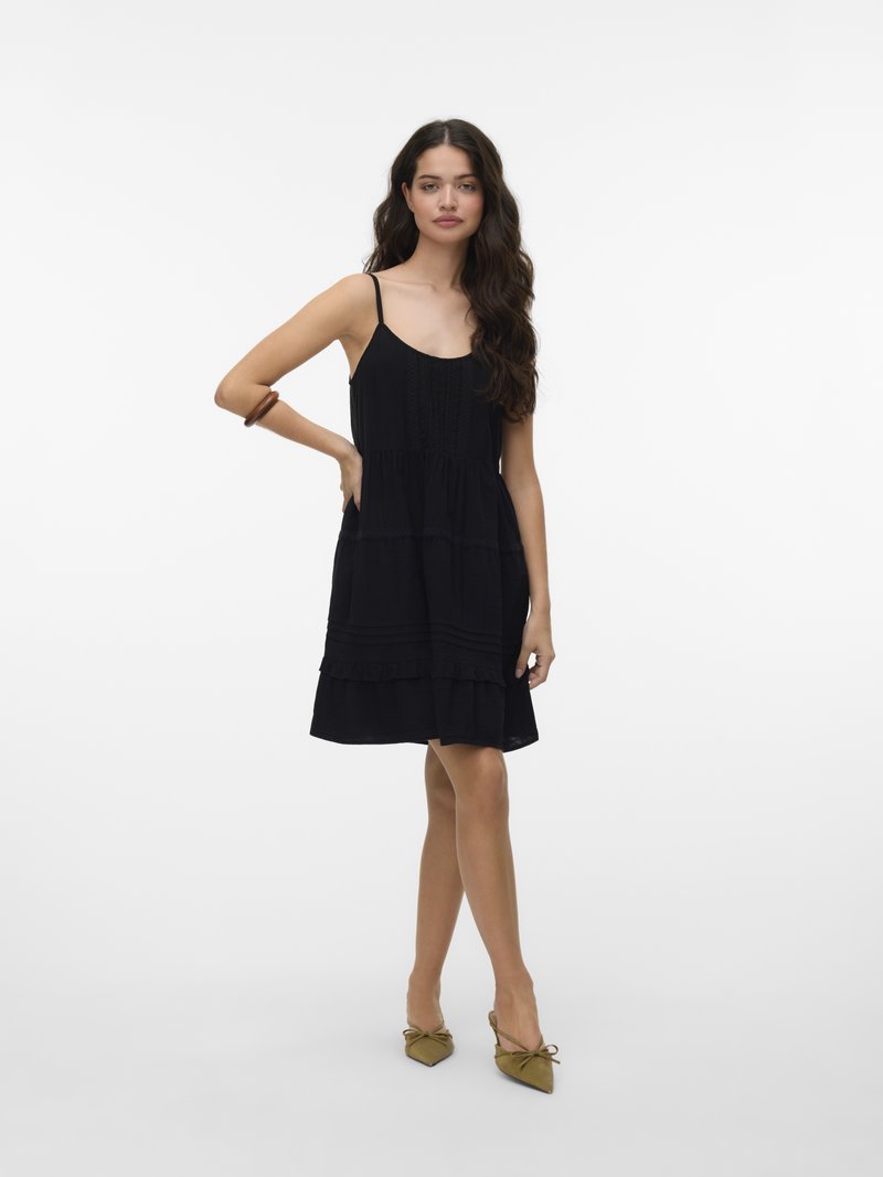 Naine seisab, üks käsi puusal, seljas must, varrukateta põlveni ulatuv kleit ja jalas oliivrohelised kikilipsudega teravaninalised kingad.