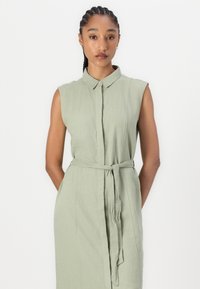 Light green sleeveless dress with a collared neck, belted waist, and a textured fabric, featuring front button closure and side pockets.