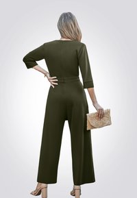 Olivgrüner Jumpsuit mit Dreiviertelärmeln, tailliertem Schnitt und weitem Bein, kombiniert mit einer strukturierten beigen Clutch.