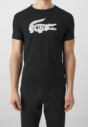Man wearing a black t-shirt with a white stylized crocodile design and "Lacoste" text across the chest.