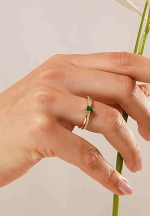 Gold ring featuring a round green stone, accented with small clear gemstones along the band, on a hand holding a flower stem.