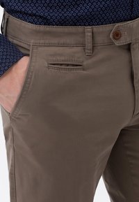 Brązowe bawełniane spodnie o dopasowanym kroju, z boczną kieszenią i brązowym zapięciem na guzik. Gładka tekstura i prosty, czysty design.