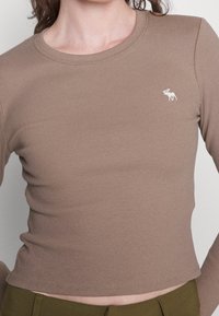 Maglietta marrone a maniche lunghe con scollo rotondo e un piccolo logo ricamato di un alce bianco. Design aderente, texture morbida e bordi senza cuciture.