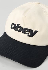 Krémovo-námorníčka baseballová čiapka s vyšitým logom "obey" v čiernej farbe, s plochým okrajom a textúrovanou tkaninou.