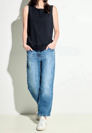 Kvinde iført sort ærmeløs top, løstsiddende blå jeans og hvide sneakers, stående med hænderne i lommerne mod en hvid baggrund.