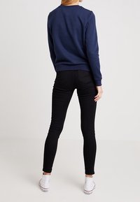 Person som bär en marinblå sweatshirt, svarta slim-fit jeans och vita högstövlar, står vänd bort från kameran mot en enkel bakgrund.