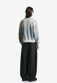 Person with shoulder-length hair wearing sunglasses, a gray gradient sweatshirt, wide black pants, and black shoes, facing away on a white background.