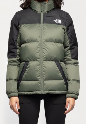 Person trägt eine grün-schwarze gesteppte Jacke mit dem The North Face Logo auf der Brust, kombiniert mit schwarzen Hosen.