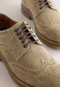 Chaussures brogues à bout golf en daim marron clair avec des lacets marron foncé et des motifs perforés détaillés sur un fond clair.