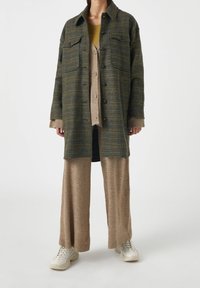 Overdimensioneret skjortejakke i grøn og blå plaid med to brystlommer, lagdelt over en beige cardigan og matchende bredbenede bukser, båret med hvide sneakers.