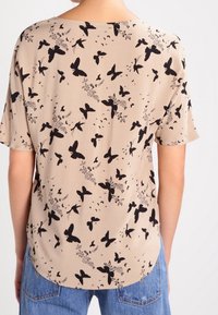 Blusa de manga corta beige con un patrón de mariposas negras y flores. Tejido suave con un escote redondo y dobladillo curvo.