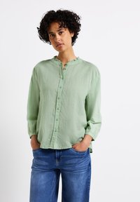 Camicetta verde chiaro con colletto arricciato, maniche lunghe e vestibilità rilassata, abbinata a jeans blu. Tessuto morbido e design casual.