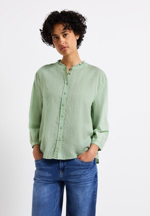 Camicetta verde chiaro con colletto arricciato, maniche lunghe e vestibilità rilassata, abbinata a jeans blu. Tessuto morbido e design casual.